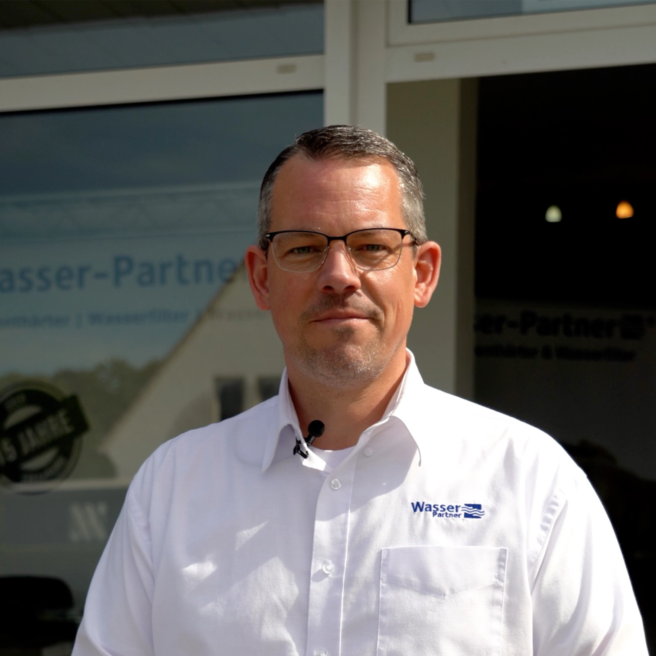 Ein Mann im weißen Hemd mit "Wasser Partner"-Logo steht vor einem Gebäude und schaut in die Kamera.