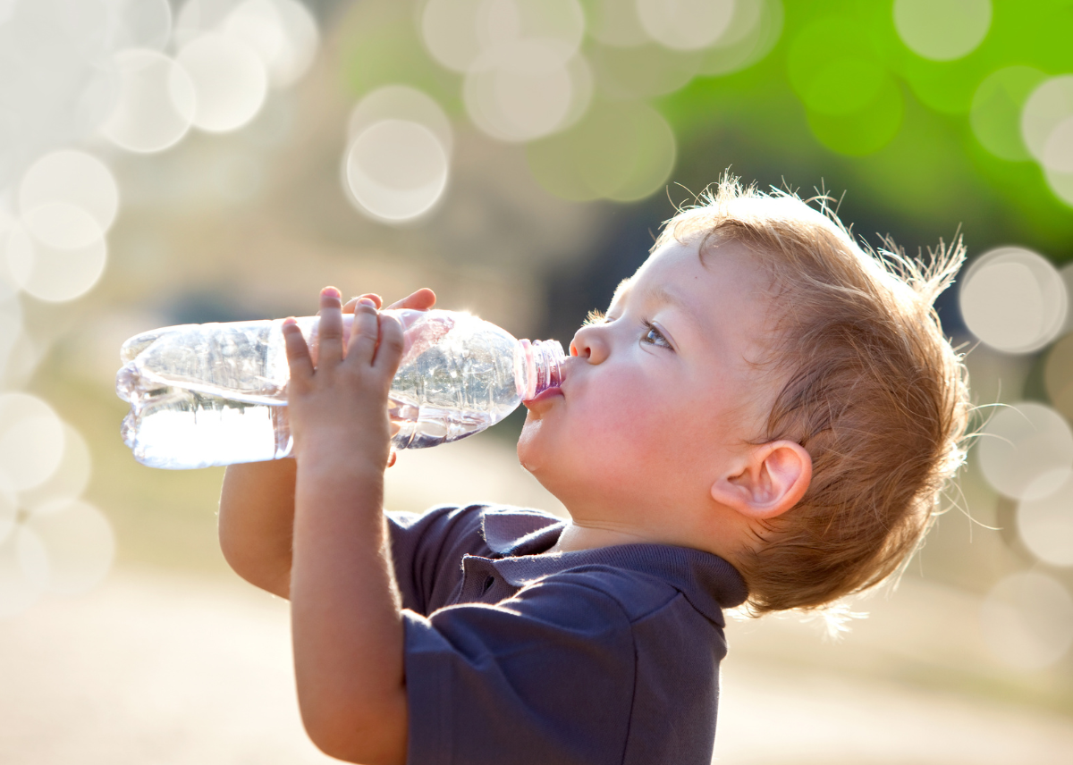 Ein Kind trinkt aus einer durchsichtigen Wasserflasche im Freien, umgeben von unscharfem Hintergrund.